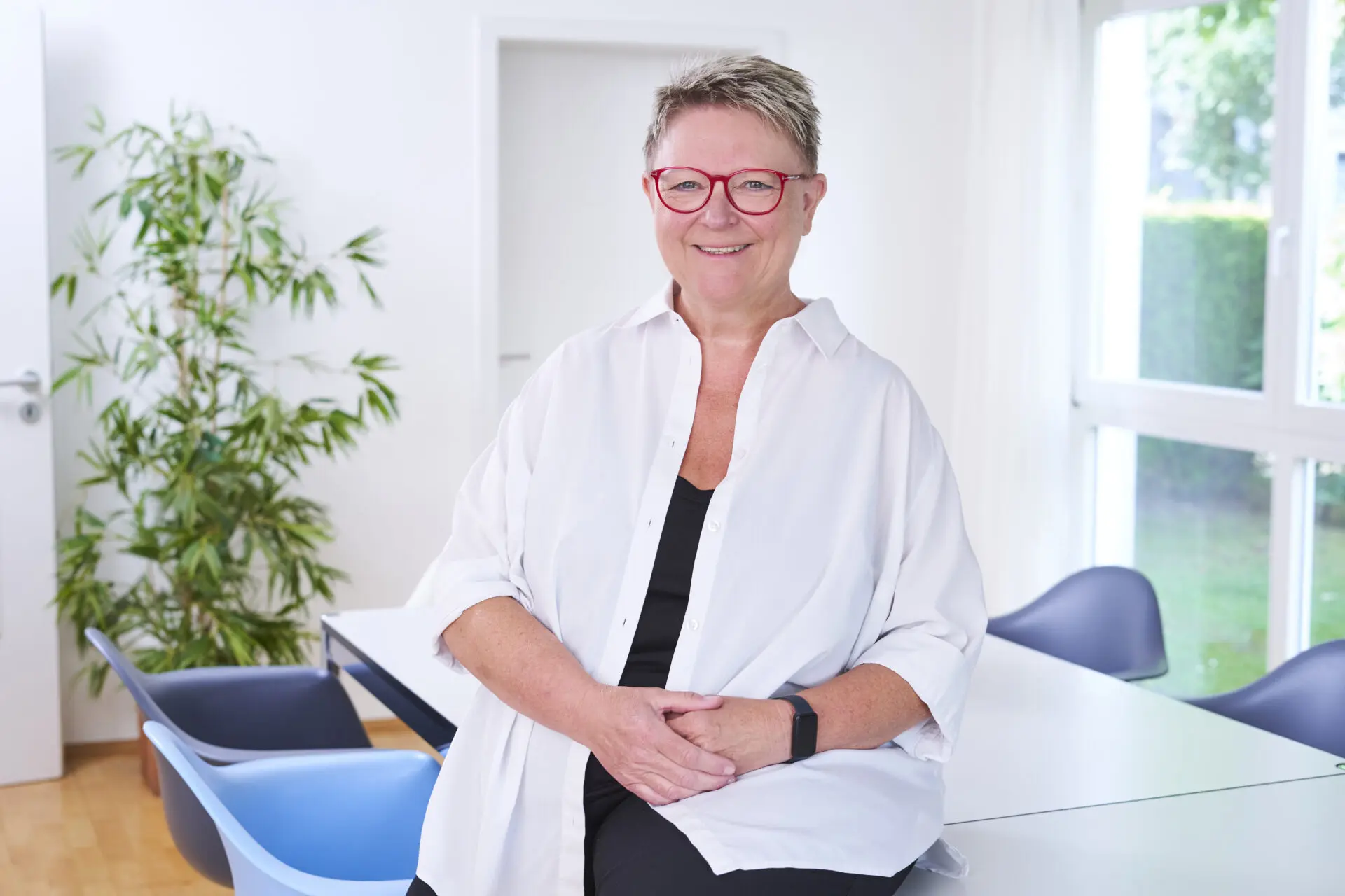 Eine Frau mit kurzen grauen Haaren und roten Brillen sitzt lächelnd an einem Tisch in einem hellen Raum.