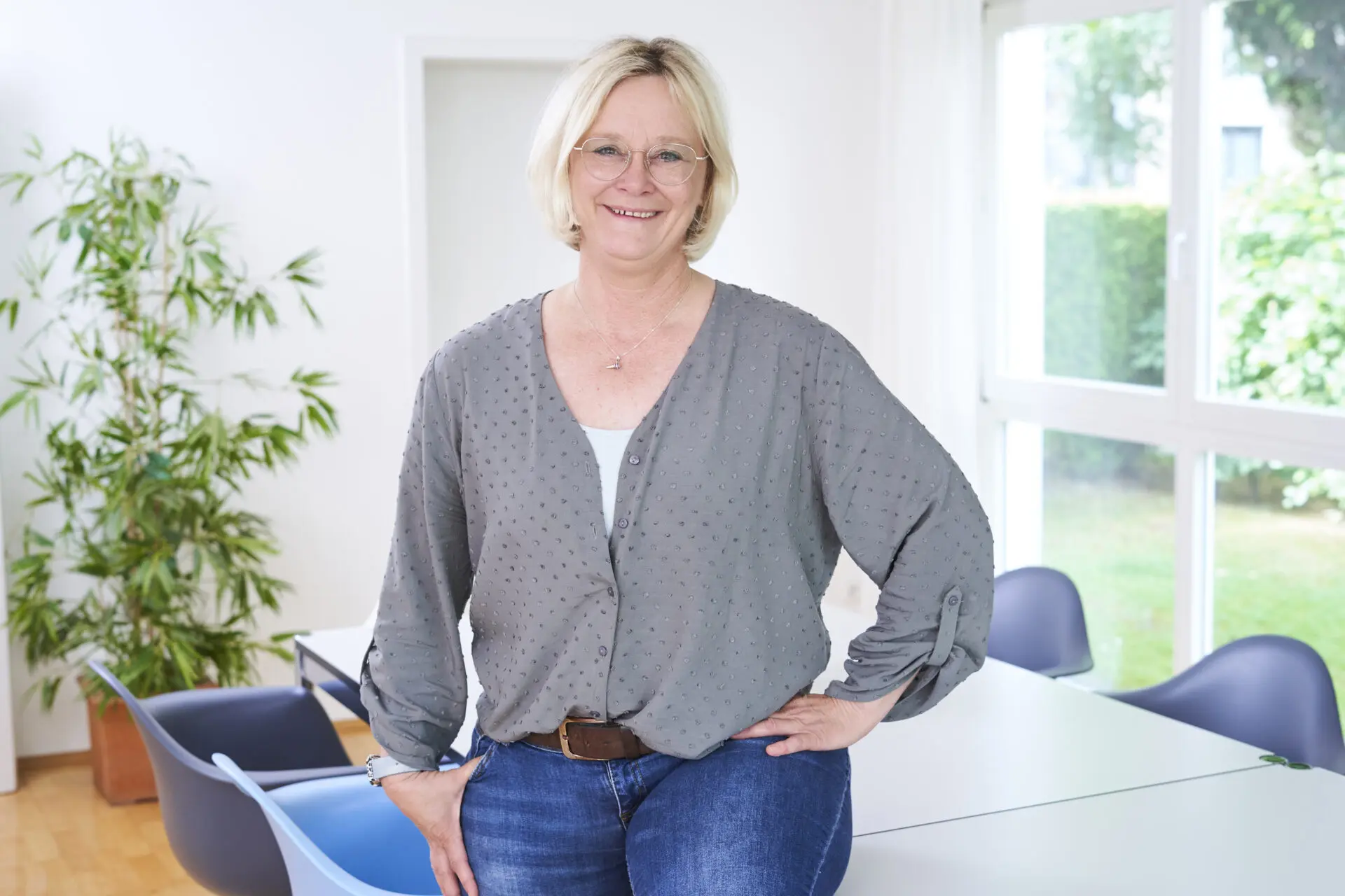 Frau mit blonden Haaren in einem grauen Hemd und Jeans steht an einem Tisch in einem hellen Raum.
