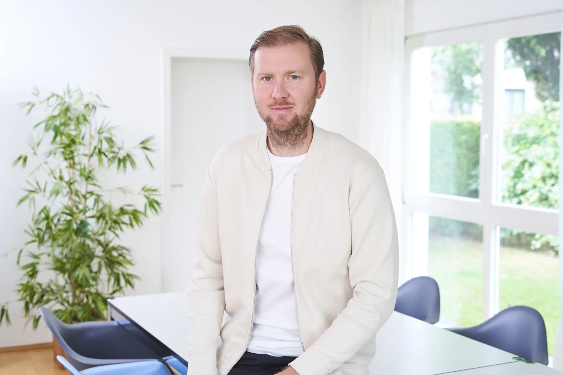 Ein Mann mit einem hellen Oberteil sitzt an einem Tisch in einem hellen Raum mit Pflanzen im Hintergrund.