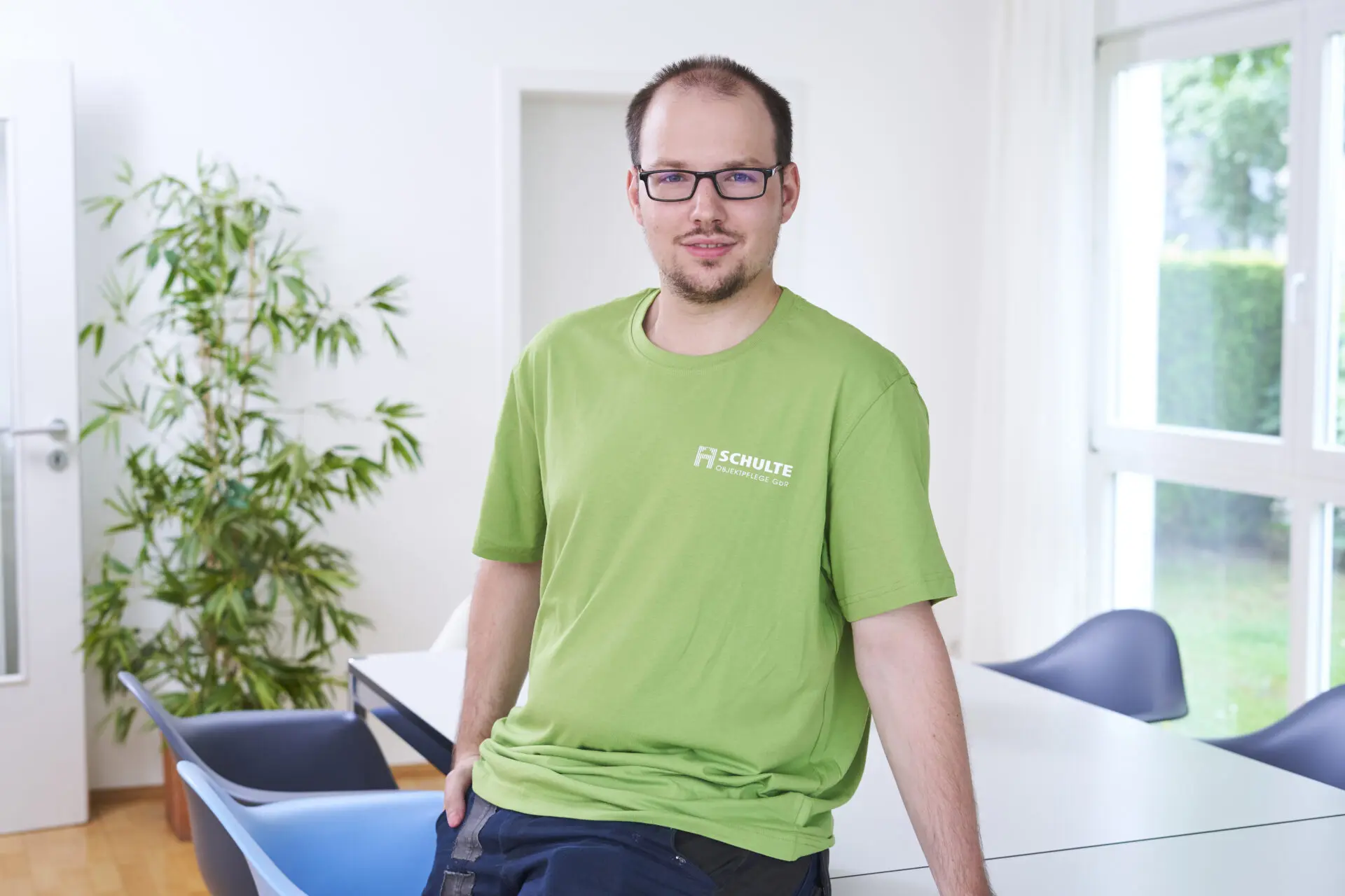 Mann mit Brille in grünem T-Shirt steht an einem Tisch in einem hellen Raum.