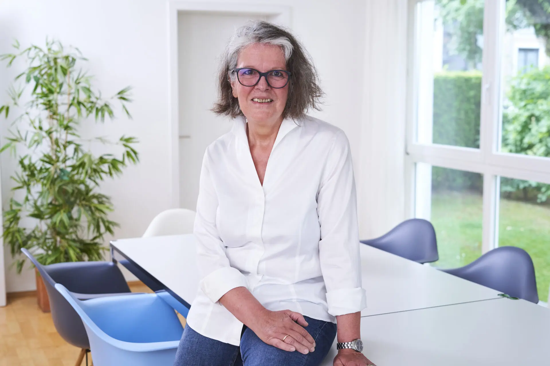 Frau mit Brille in einem weißen Hemd sitzt auf einem Tisch in einem hellen Raum.