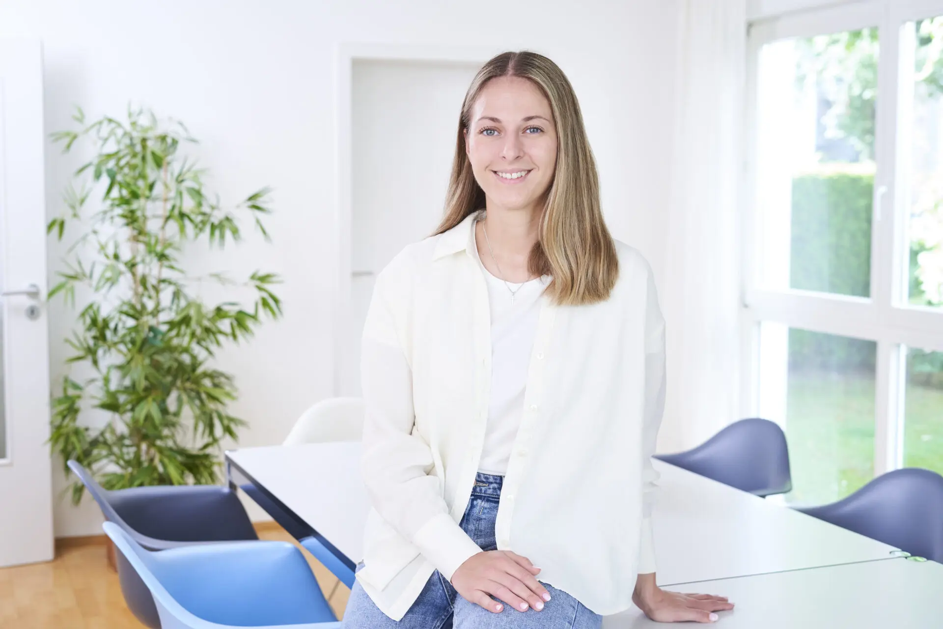 Junge Frau mit langen Haaren, die in einem hellen Raum an einem Tisch steht und lächelt.