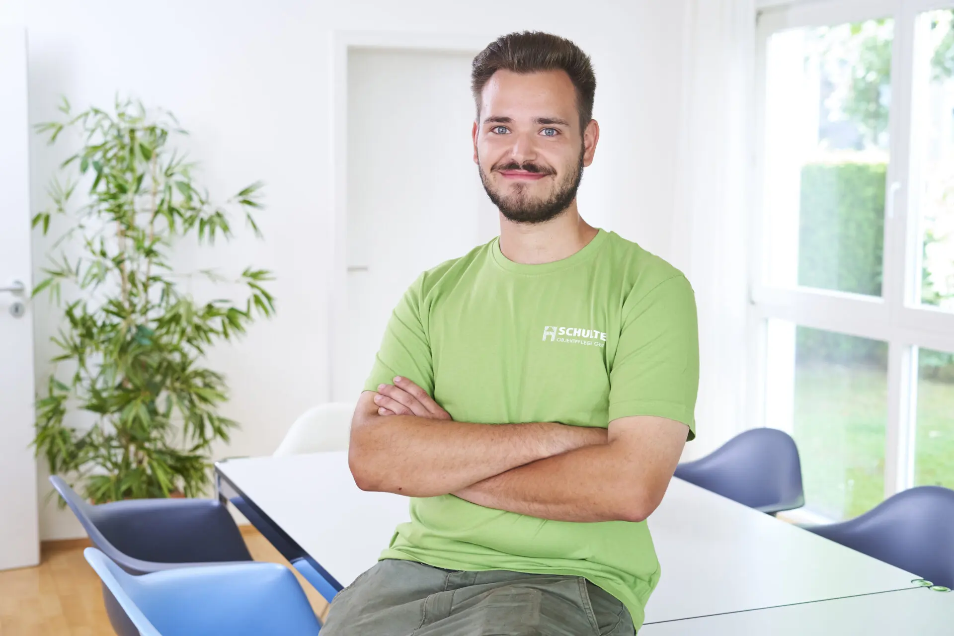 Junger Mann mit grünem T-Shirt und verschränkten Armen, lächelnd in einem Raum mit Tisch und Stühlen.