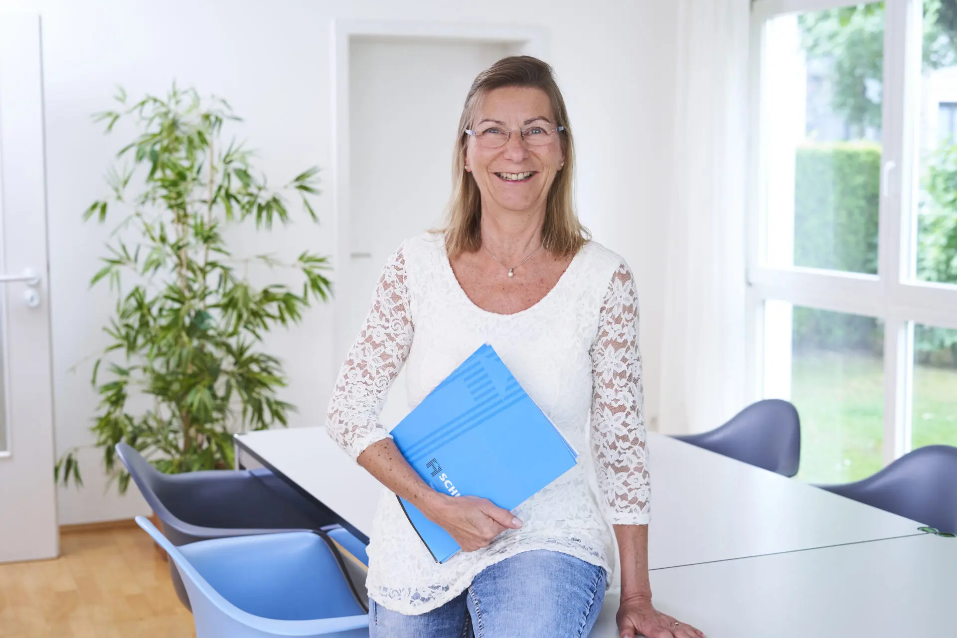 Eine Frau mit Brille steht in einem Büro und hält eine blaue Mappe in der Hand.