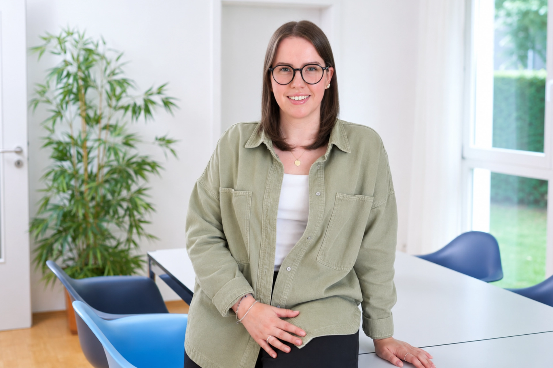 Hannah Vernhold sitzt lächelnd in einem hellen Büro mit Brille und olivgrünem Hemd, passend zum individuellen Teamauftritt von Immobilien Schulte.