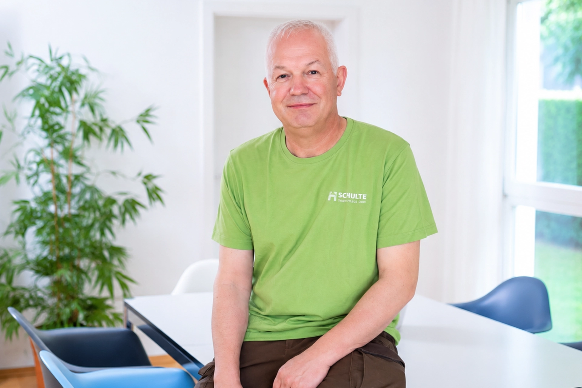 Markus Klar sitzt in einem hellen Besprechungsraum und trägt ein grünes Schulte-T-Shirt, während im Hintergrund Pflanzen und große Fenster zu sehen sind.