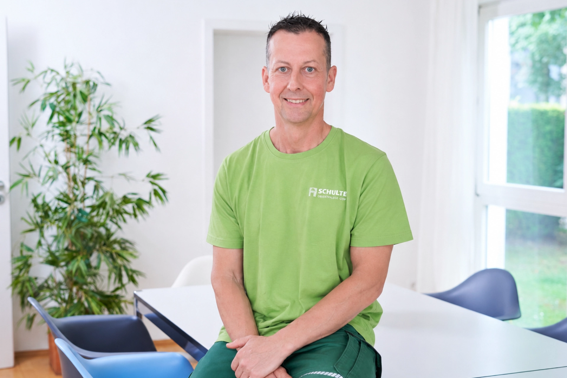 Peter Rocholl sitzt in einem hellen Besprechungsraum auf einem Tischrand, trägt ein grünes Schulte-T-Shirt und blickt freundlich in die Kamera.